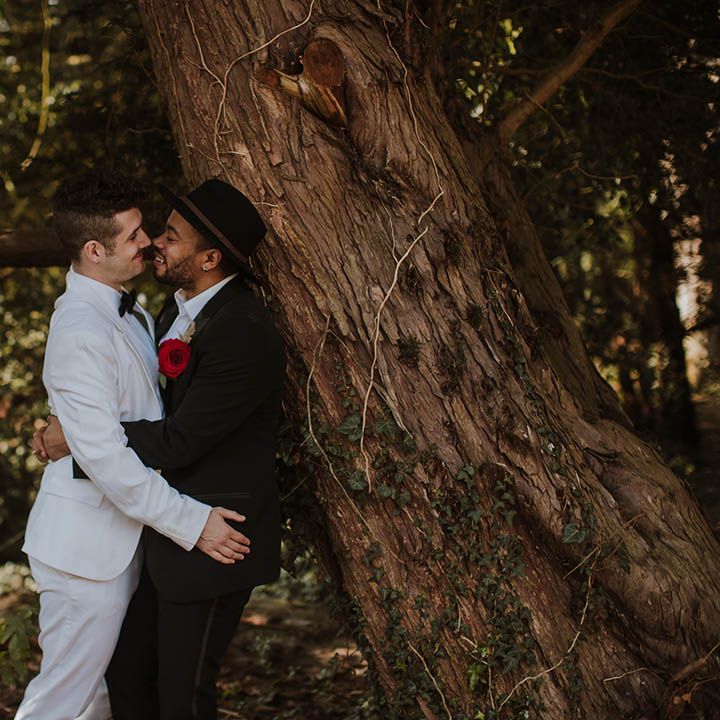 Groom in a white suit embracing his partner in a black groom suit with red Gerbra buttonhole flowers 
