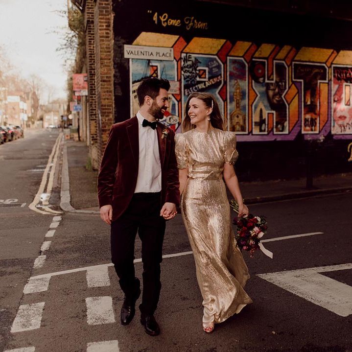 Bride in puff sleeve gold wedding dress and bow detail holds hands with groom in burgundy velvet suit jacket