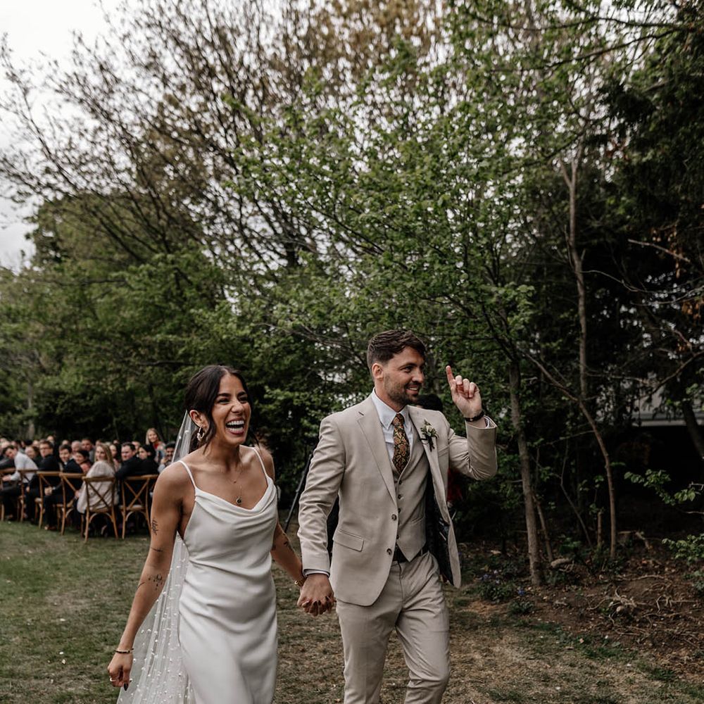 Groom in a stone coloured suit holding hands with his bride in a satin slip wedding dress and pearl veil 