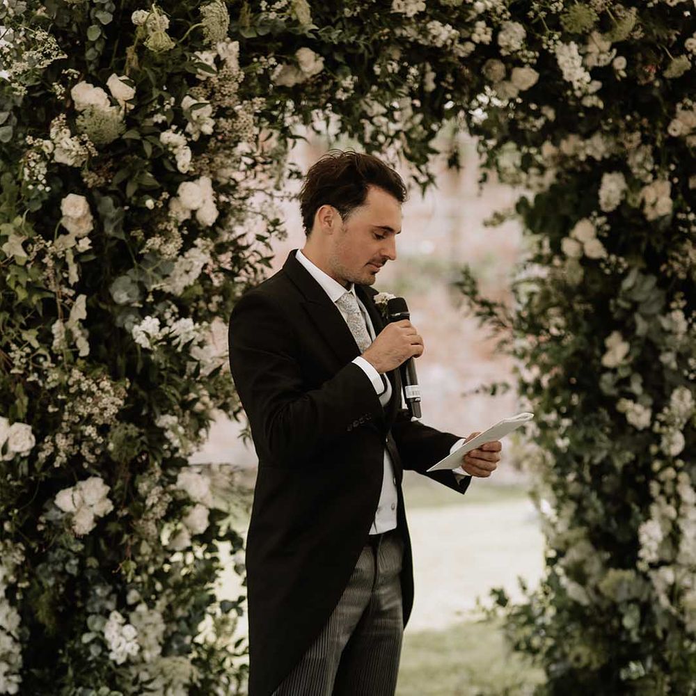 Groomsmen in morning suit and striped trousers performs a wedding reading for outdoor marquee wedding next to white flower arch