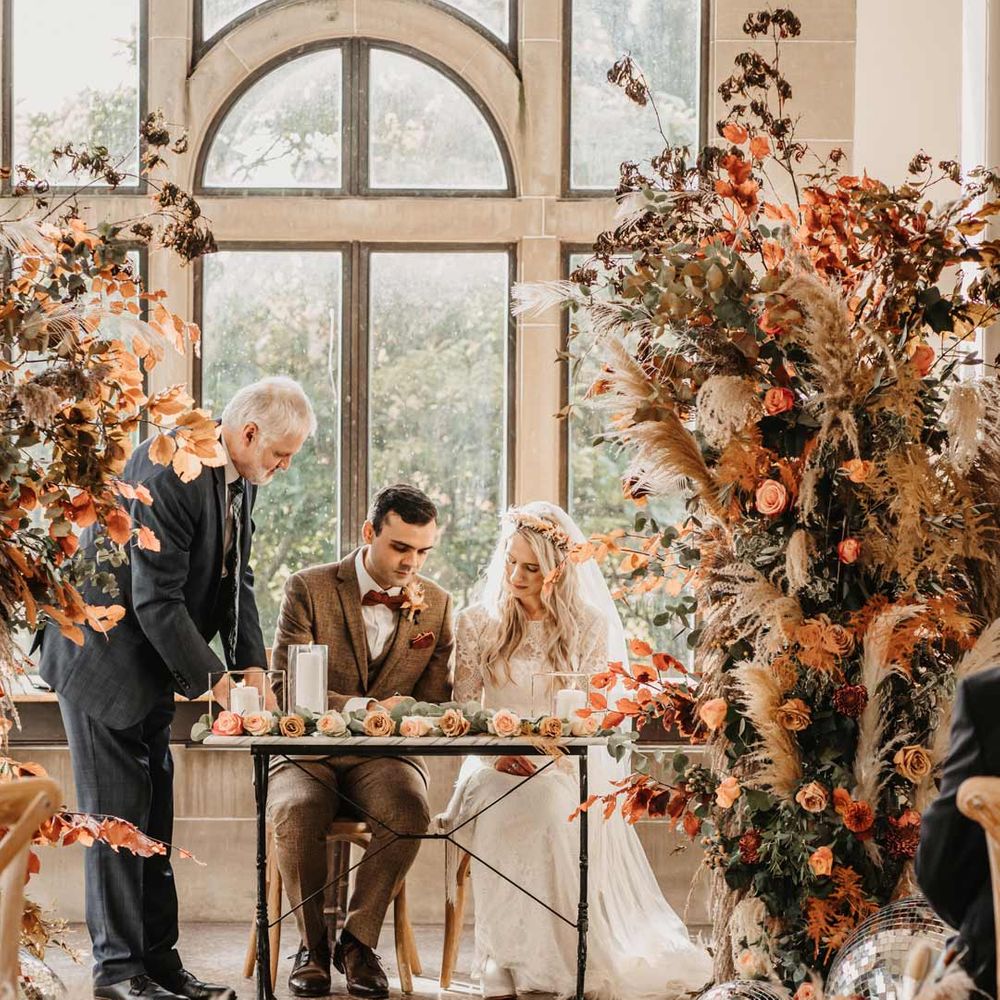 Bride in lace dress and flower crown and groom in orange themed wedding singing the register