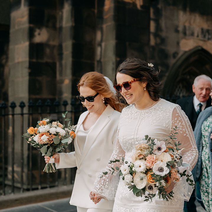Brides walk holding hands whilst wearing sunglasses and holding colour floral bouquets 