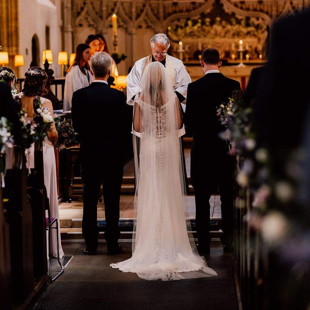 Bride in sheer back lace wedding dress stands inbetween the groom and her father as they stand at the altar for their church wedding ceremony 