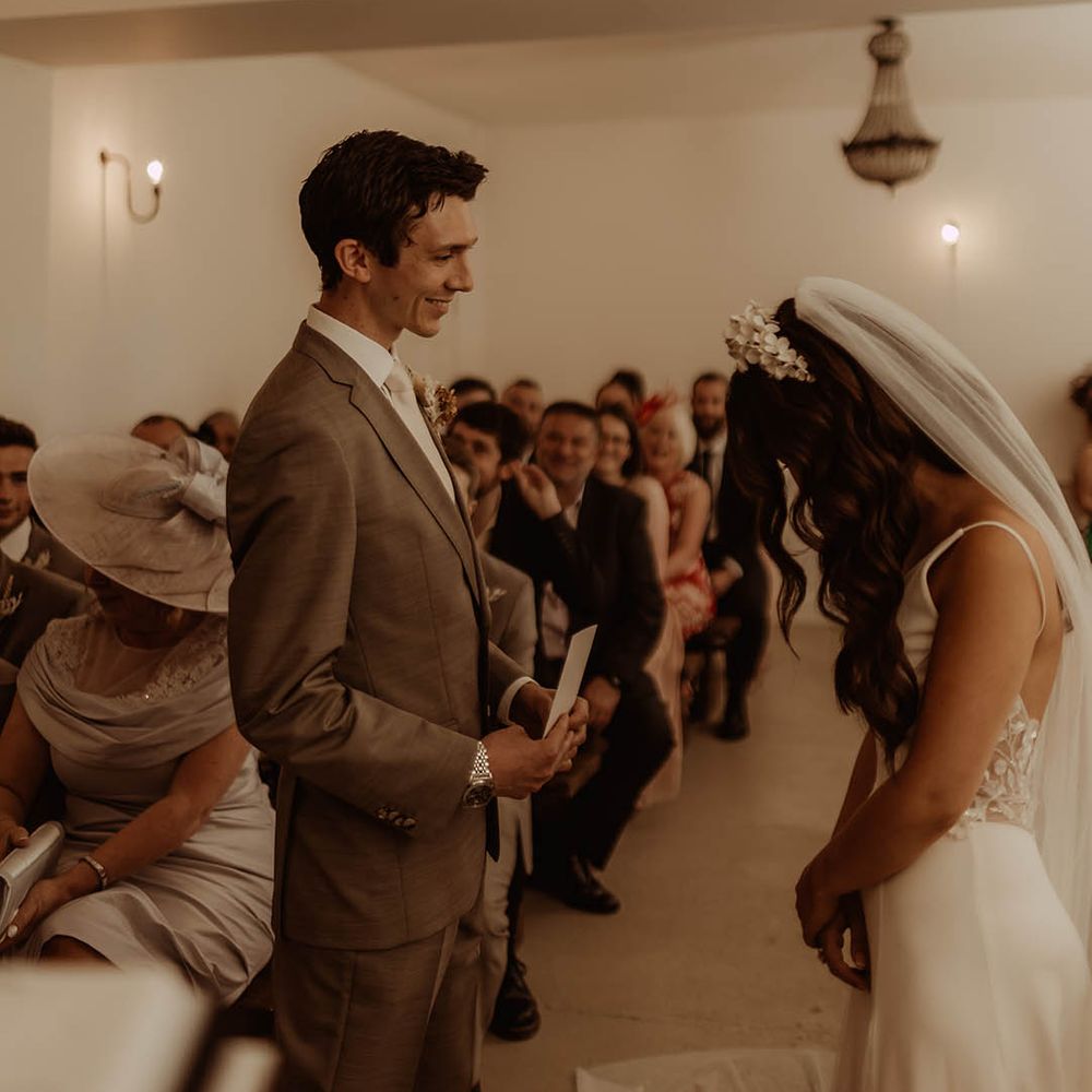 Groom smiles at the bride in neutral three piece suit as he reads out his wedding vows to the bride 