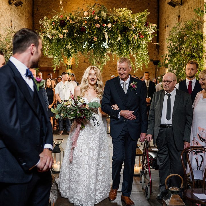Father of the bride in blue suit and pale blue tie walks the bride down the aisle in a strapless lace wedding dress 
