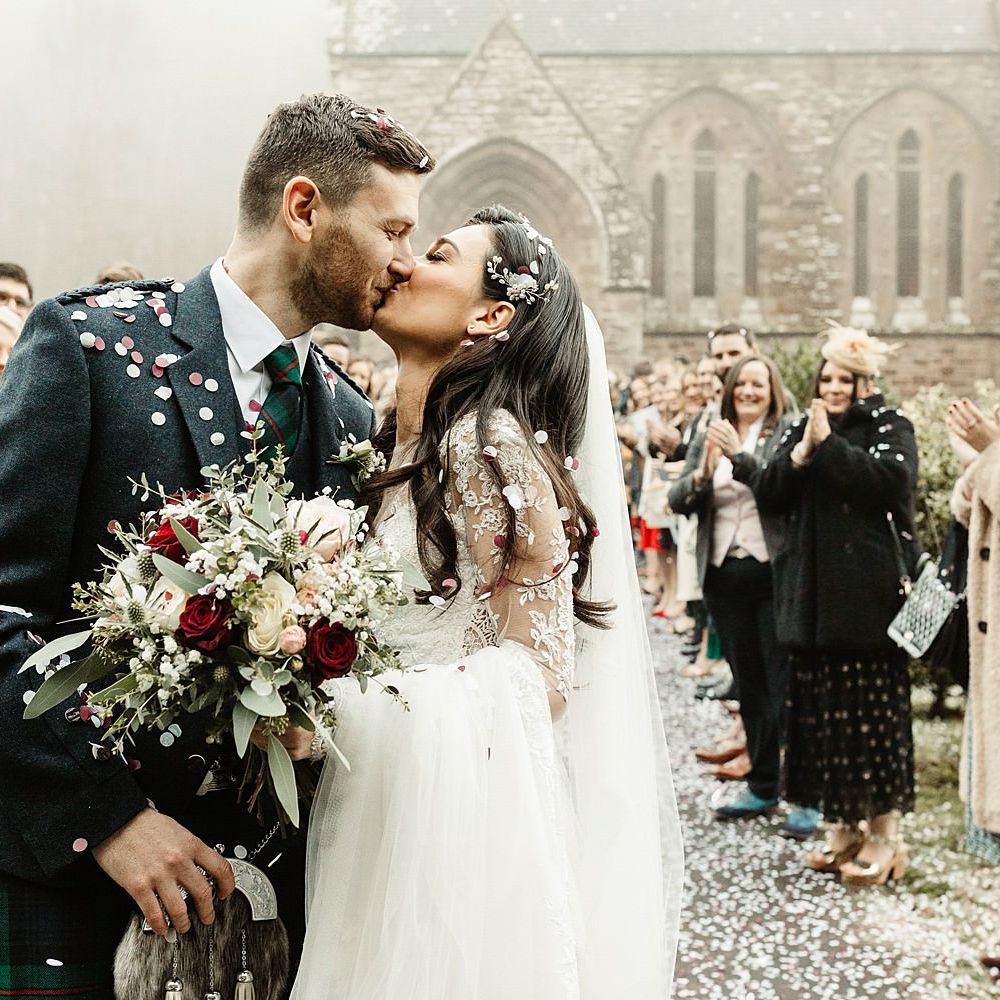 Bride & groom kiss outside church on winter wedding day surrounded by white and red confetti as bride holds matching coloured bouquet 