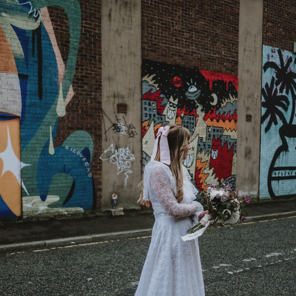 Bride wearing vintage lace wedding dress holds pink and white floral bouquet whilst standing in front of brightly coloured brick wall 