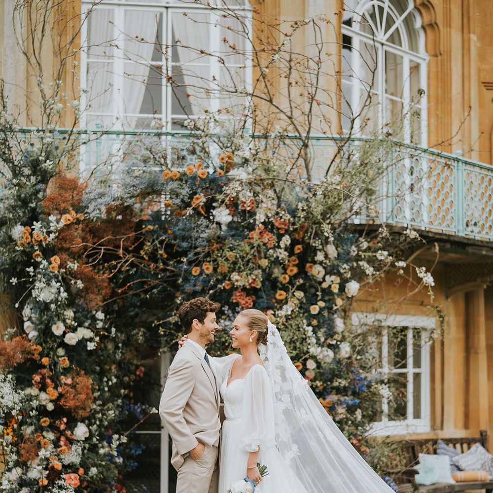 Groom in a beige suit standing with the bride in a white wedding dress and flower veil in front of colourful flower arch 