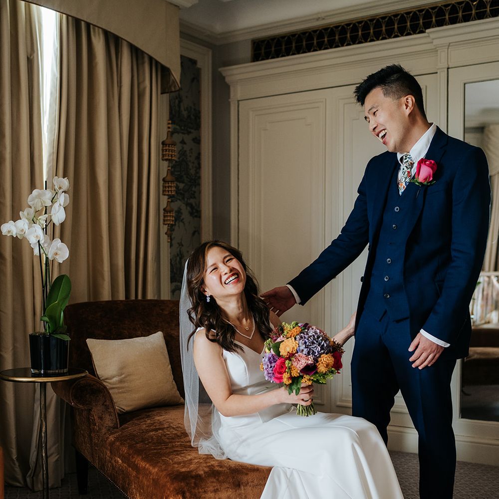 Bride holds brightly coloured floral bouquet and sits beside her groom in three piece suit with pink rose buttonhole 