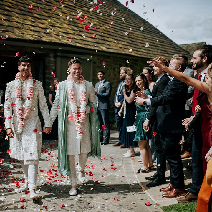 Grooms walk hand in hand through colourful petal confetti after barn wedding ceremony 