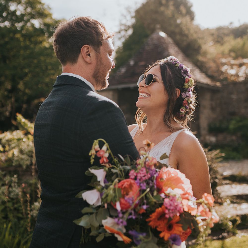 Bride wearing sunglasses and hugs her groom whilst holding colourful floral bouquet 