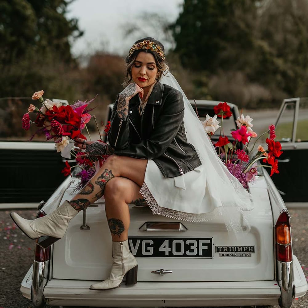 Bride sitting on the bonnet of off white classic convertible wedding car wearing blazer style wrap around short wedding dress with delicate fringe detailing and black leather jacket