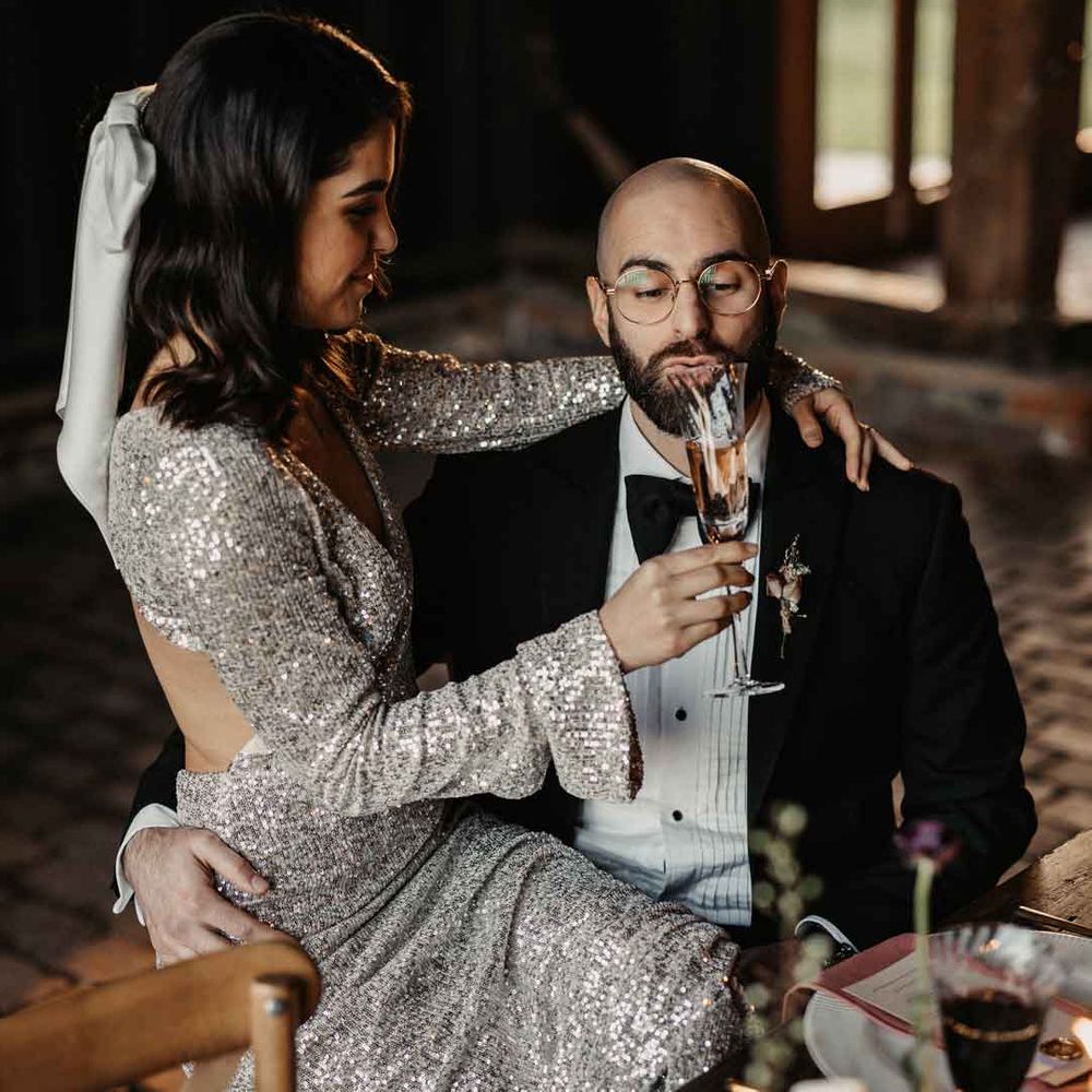 Bride in open back long sleeve silver sparkly wedding dress giving groom in classic black tuxedo and black bowtie a glass of champagne 