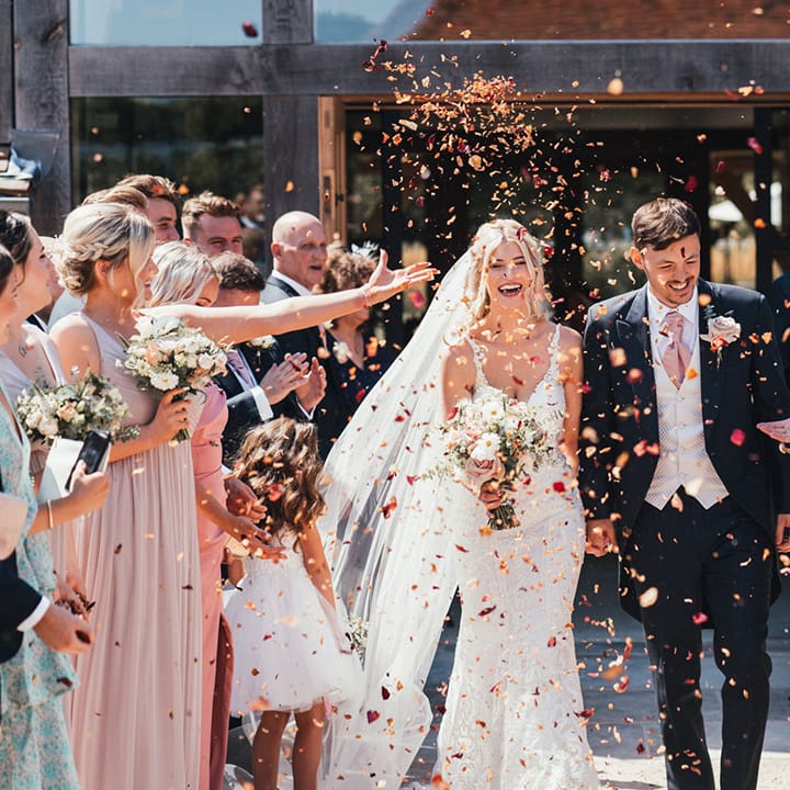 Groom in three piece suit with pink tie walking with the bride in a fitted lace wedding dress walking along as confetti is thrown over them 