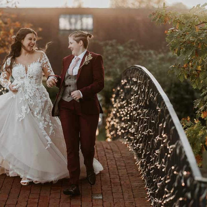 Bride in long sleeve lace illusion sleeve wedding dress with overlay walking over a bridge at Bassmead Manor Barns with bride in maroon bridal suit with grey waistcoat, dusky pink tie and autumnal dried flower boutonniere 