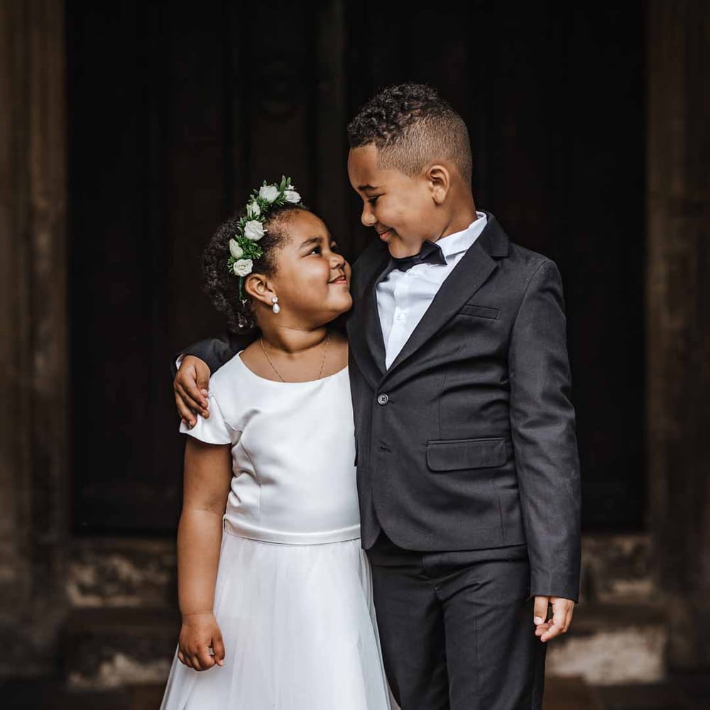 Flower girl wearing short sleeve satin dress and flower crown looking up at page boy in black suit and bowtie at Lains Barn wedding