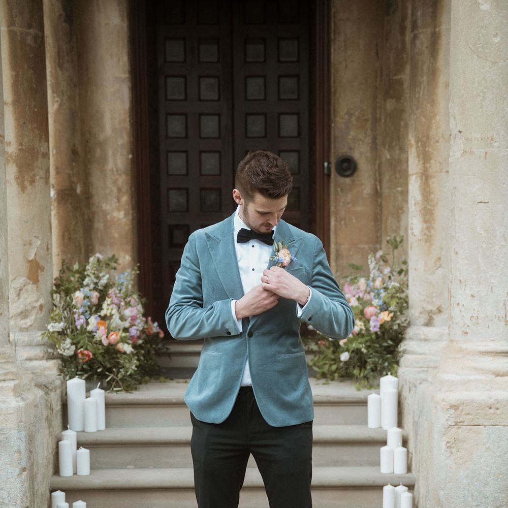 Groom in black trousers, black bow tie, blue velvet blazer and pastel floral buttonhole 