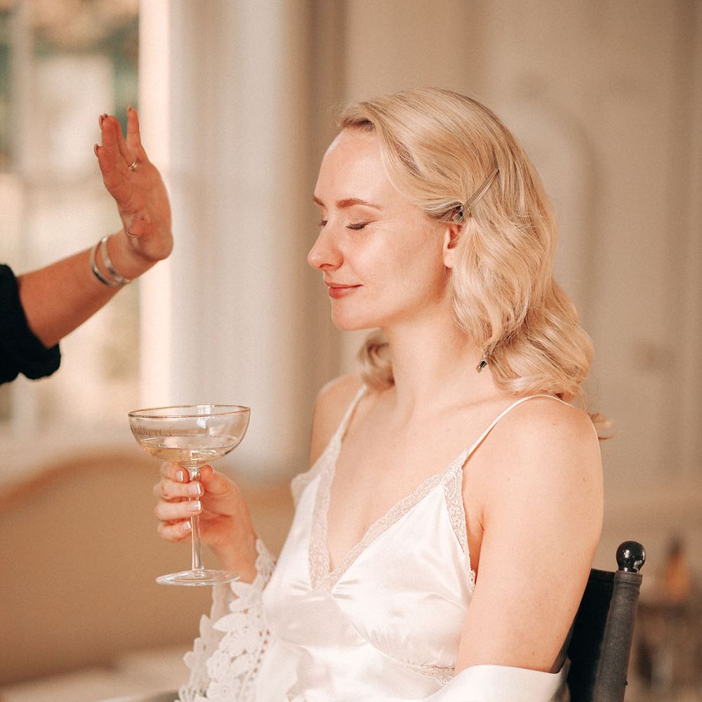 Bride in white satin pyjamas sits getting her makeup done for the wedding day 