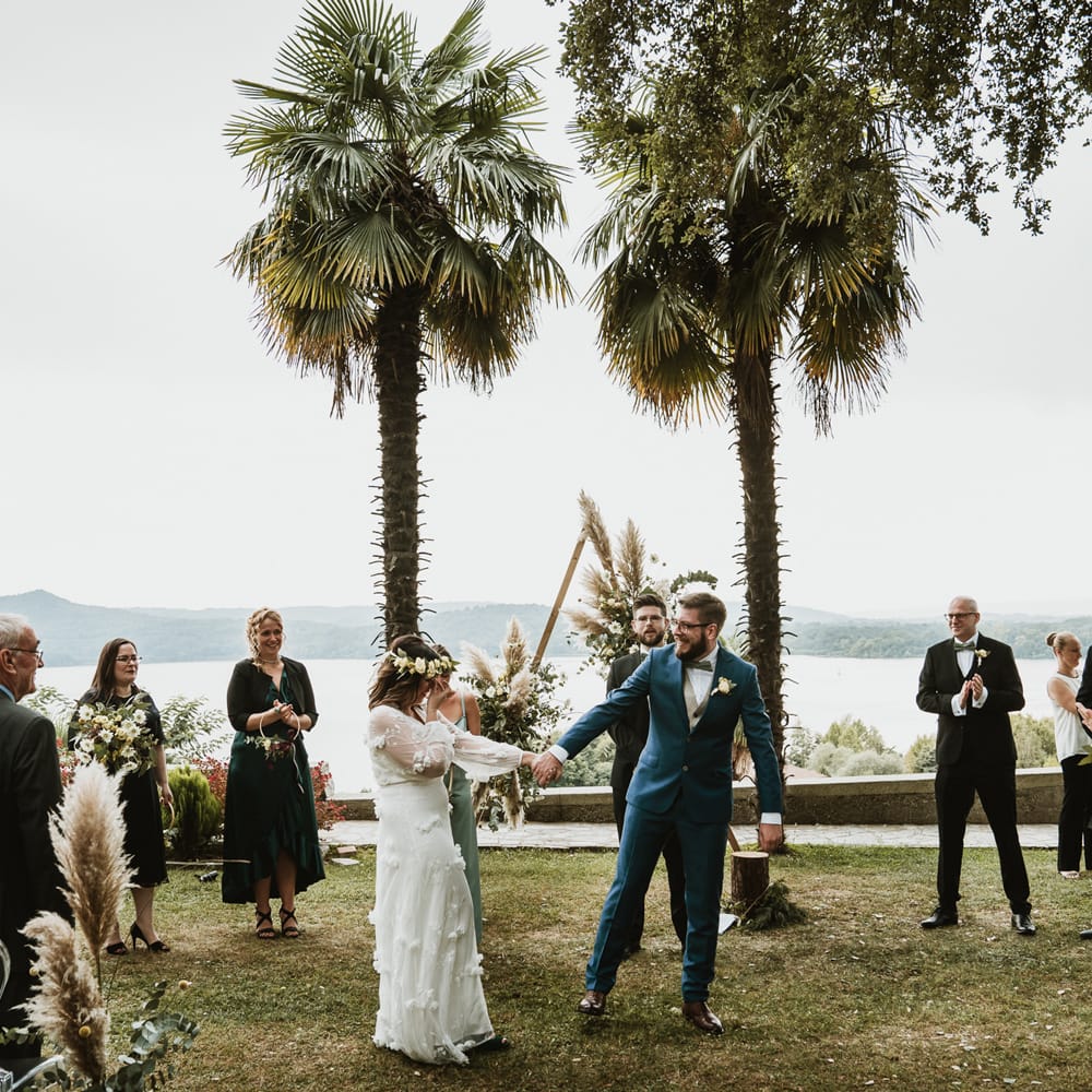 Bride in a white wedding dress with flowers holding hands with her groom in navy blue suit and bow tie at their outdoor hand fasting ceremony 