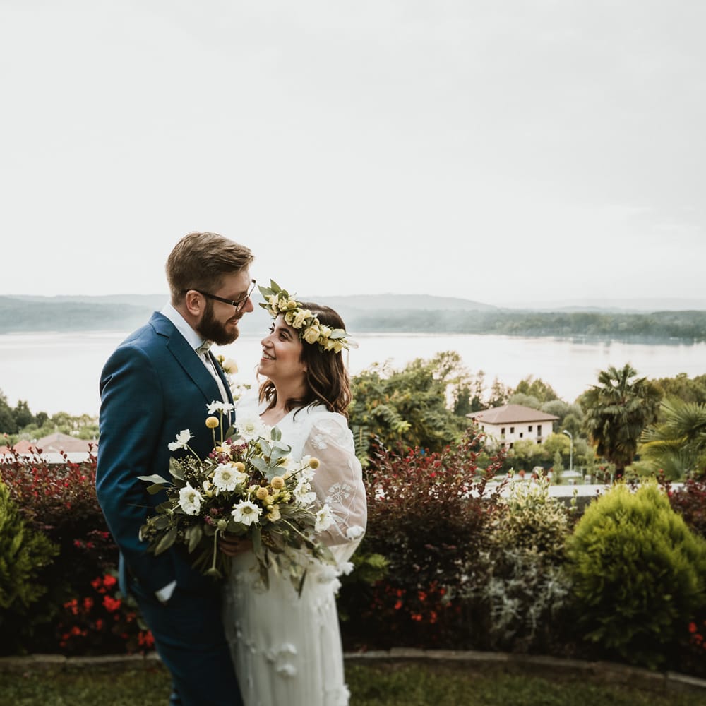 Portrait of the boho bride in a white wedding dress with flowers smiling at her groom in a blue and green suit with Lake Viverone, Italy in the background