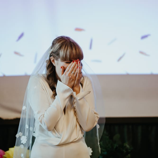 Stylish bride in a wrap jumpsuit with button detail on the sleeves covering her face with excitement 