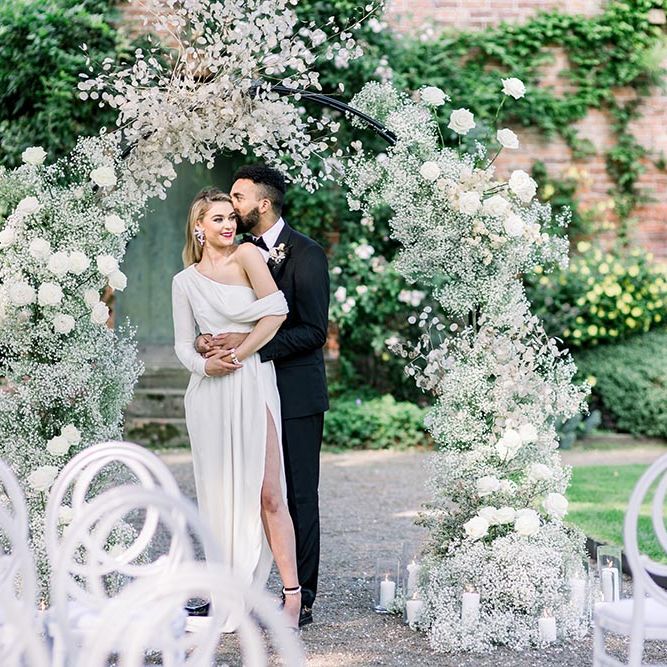 Garthmyl Hall wedding inspiration with groom in a tuxedo kissing his bride in a wrap dress with one shoulder under a white flower arch 