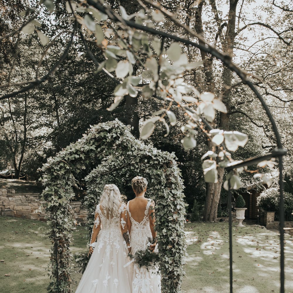 Two brides walk under ivy covered arches at Crab & Lobster fairytale wedding. 
