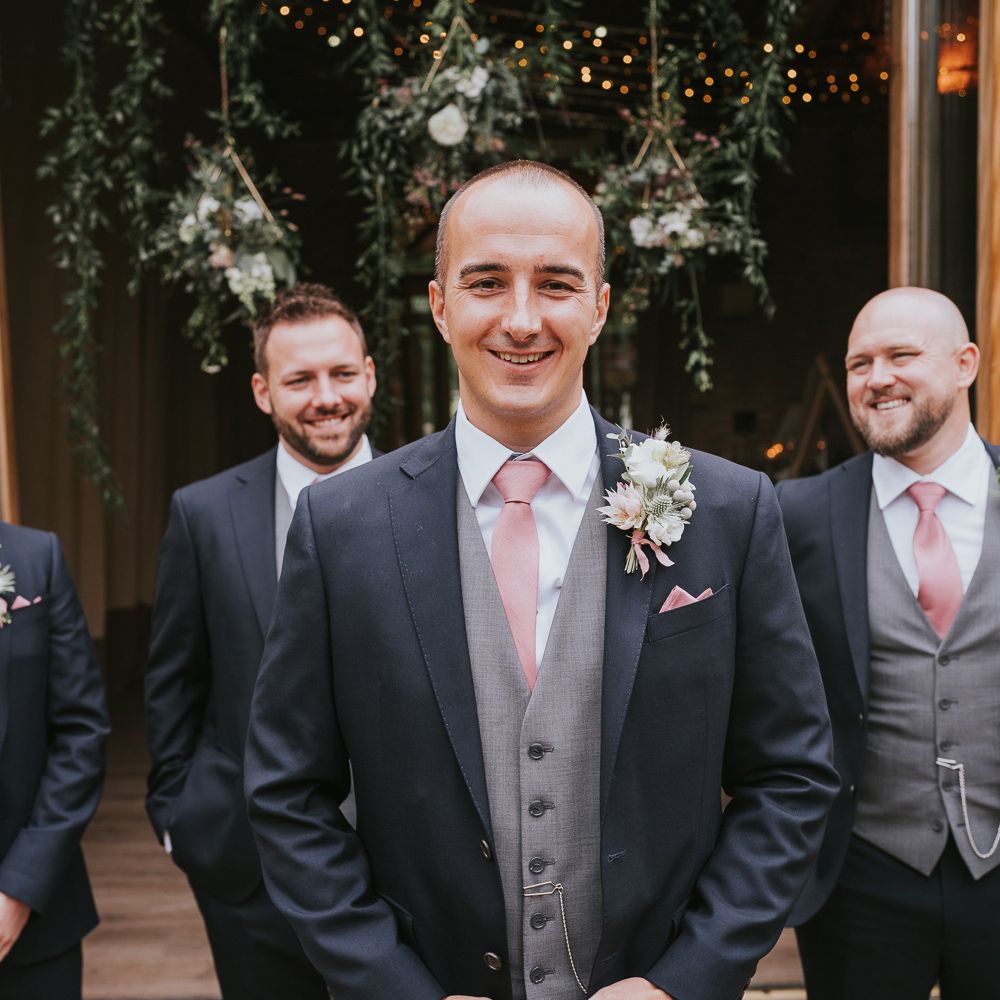 Groom and groomsmen in dark three piece suits with grey waistcoats, pink ties and protea and thistle buttonholes at Tythe Barn wedding with barn wedding flowers