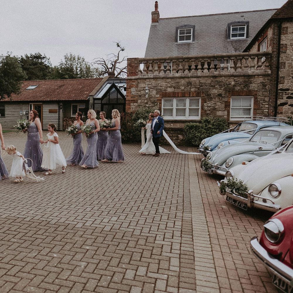 Bridesmaids in blue beaded cami bridesmaids dresses and flower girls in white dresses walk across courtyard followed by bride in Grace Loves Lace wedding dress and veil arm in arm with father in blue suit at Isle of Wight wedding