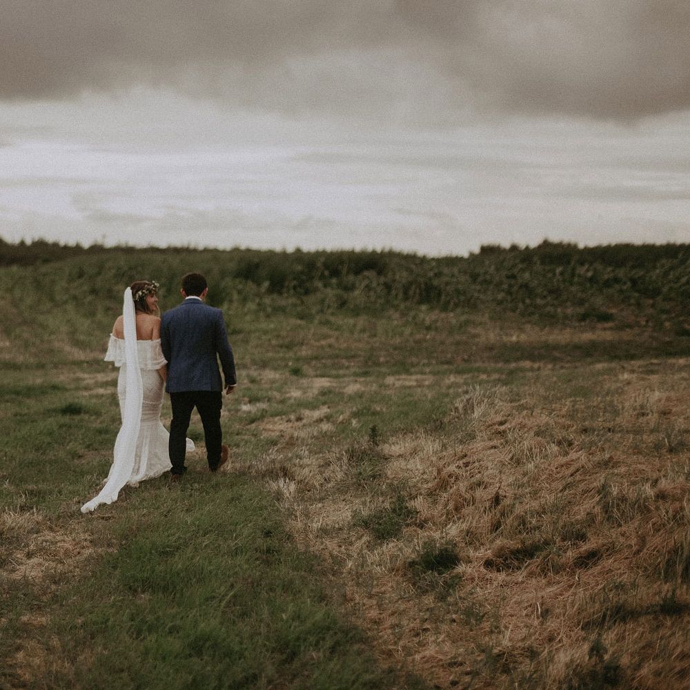 Bride in Grace Loves Lace bardot wedding dress, veil and flower crown walks arm in arm with groom in blue suit jacket walk through field at Isle of Wight wedding with macrame wedding decor