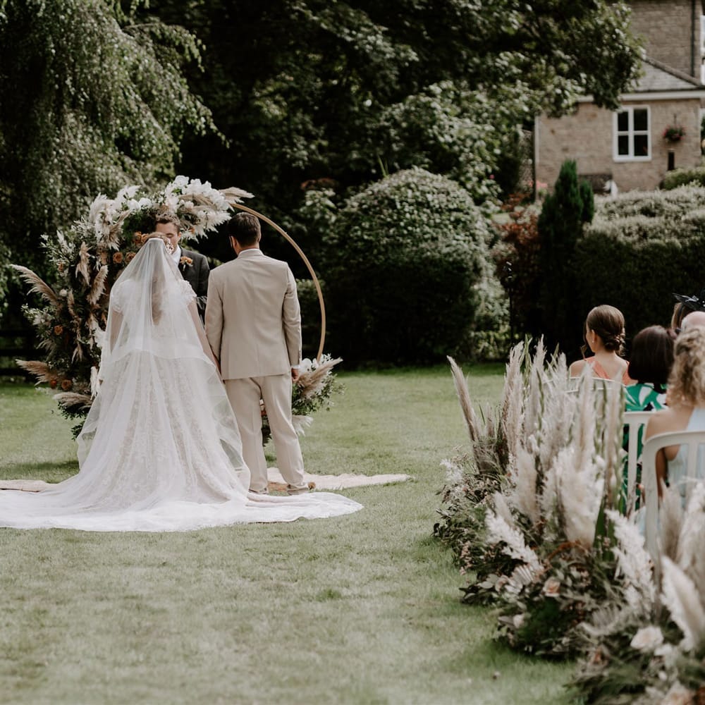 Bride in homemade wedding dress and veil stands at floral Moorgate altar with groom in beige suit during DIY garden wedding ceremony in Bedfordshire