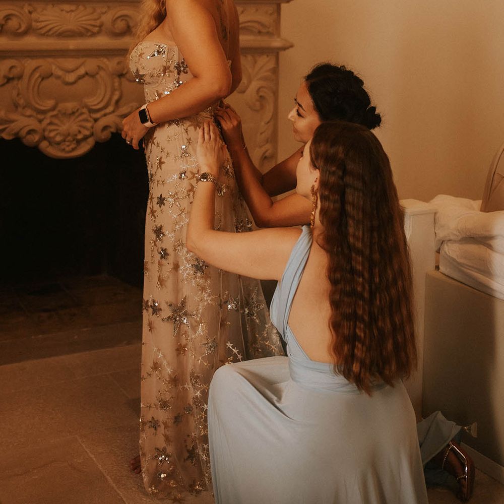 Bridesmaids kneel behind bride whilst doing up her wedding gown