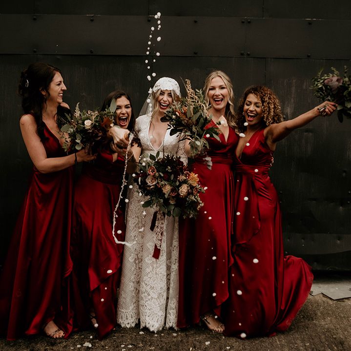 Fun bridal party portrait with bride in a lace wedding dress and bridal party in satin red bridesmaid dresses spraying champagne 