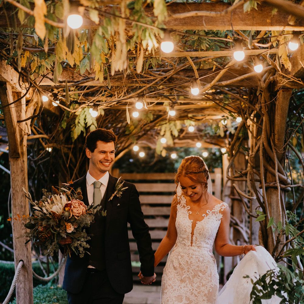 Bride in deep v lace wedding dress holding tulle train walks under Edison bulb canopy with groom in sage green tie holding mixed bridal bouquet