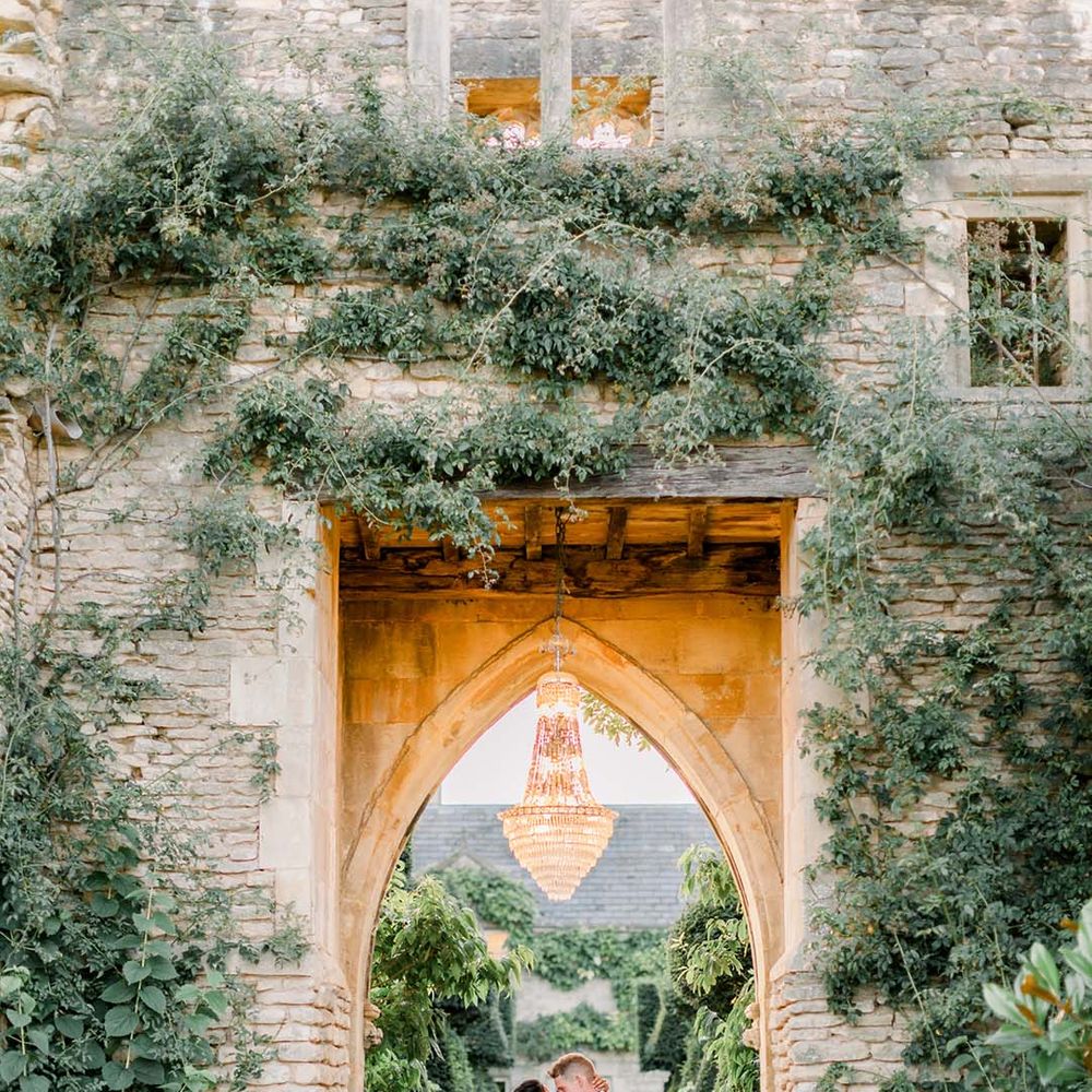Bride & groom kiss under archway outdoors with chandelier hanging above them on their wedding day