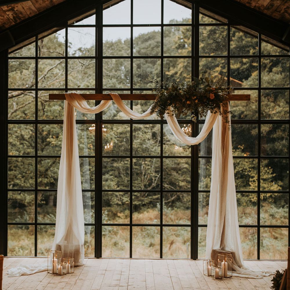Wooden wedding arch with white draping, floral cloud and candles in front of large window at the Hidden River Barn wedding venue