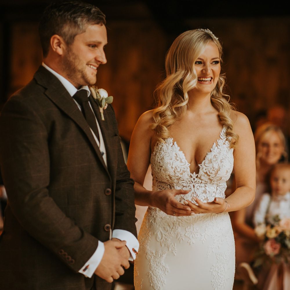 Smiling bride with curled hair in lace wedding dress stands with groom in brown three piece suit and buttonhole during rustic barn wedding ceremony