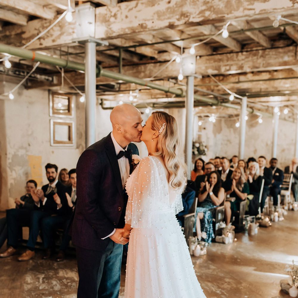 Groom in navy tweed suit with brown shoes kisses bride in pearl wedding dress with train during wedding ceremony at Holmes Mill
