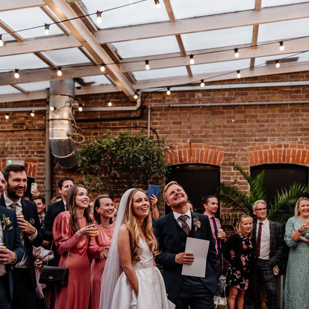 Groom in a navy suit and bride in a princess wedding dress with pockets listening to their wedding speeches 
