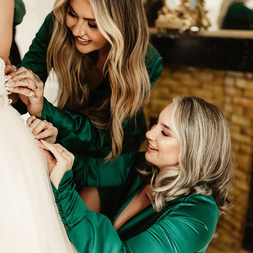 Bridesmaids in emerald green long sleeve dresses and French tip nails do up buttons on the bride's wedding dress