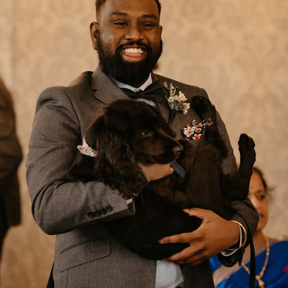 Groom in dark grey suit and white neutral boutonniere holding black puppy 