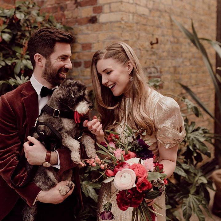 Groom in burgundy suit jacket with bride in gold wedding dress holding their pet dog wearing red bow tie collar 
