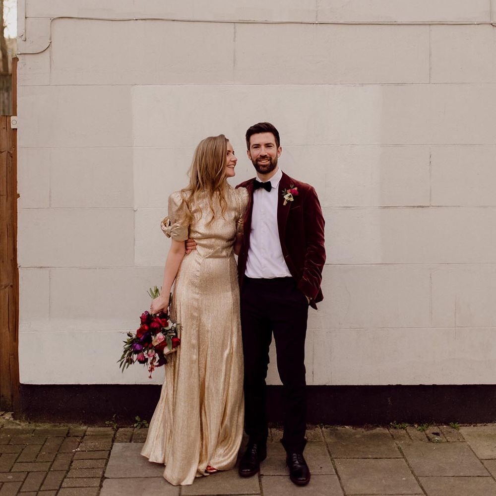 Groom wearing black tie and red velvet suit jacket has his arm around the bride in sparkly gold wedding dress holding dark wedding bouquet