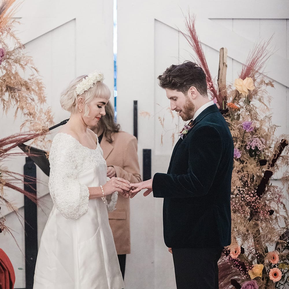 Bride in short wedding dress with white flower headband places the ring on the groom's finger at wedding ceremony