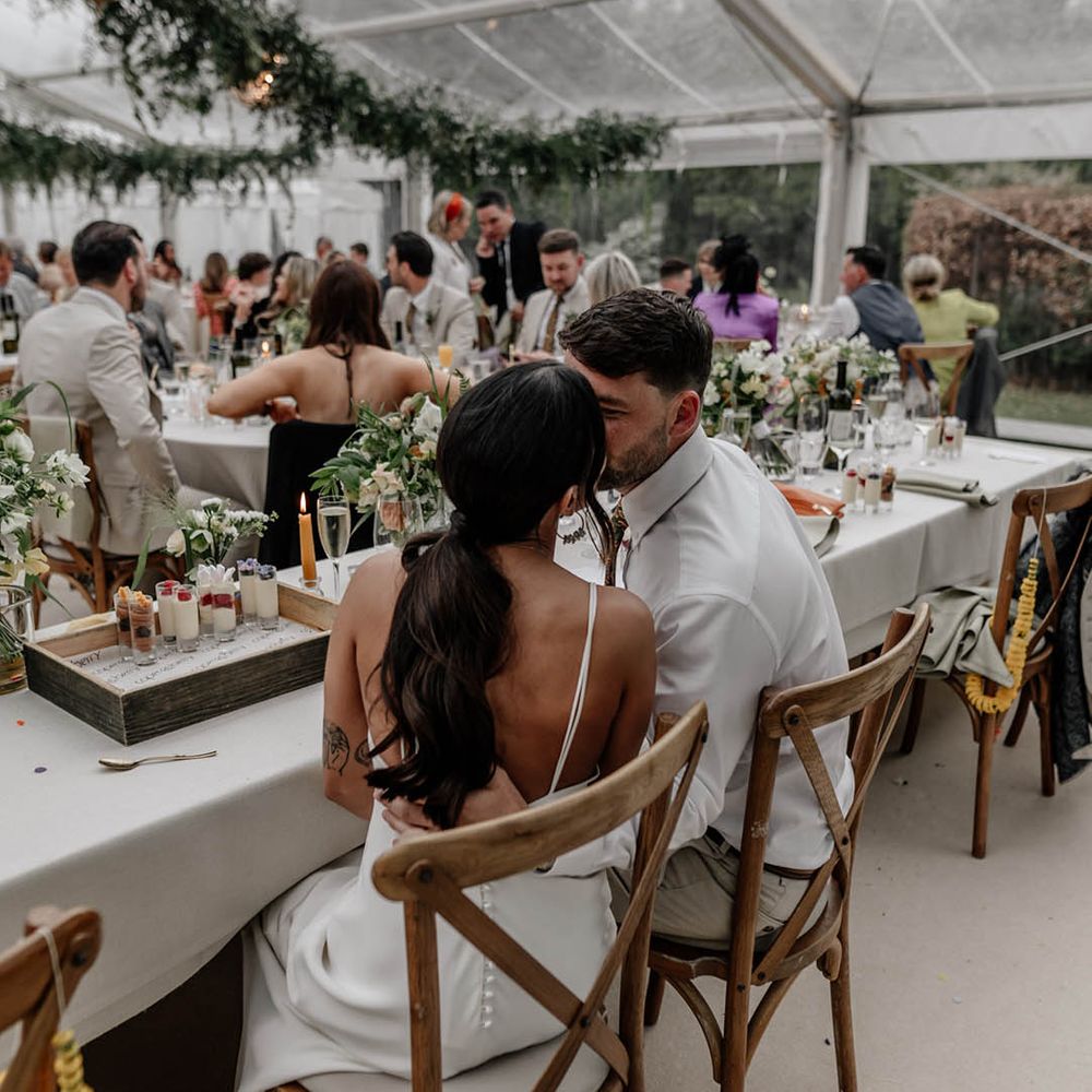 Bride and groom kissing at the top table of their clear glass marquee reception 