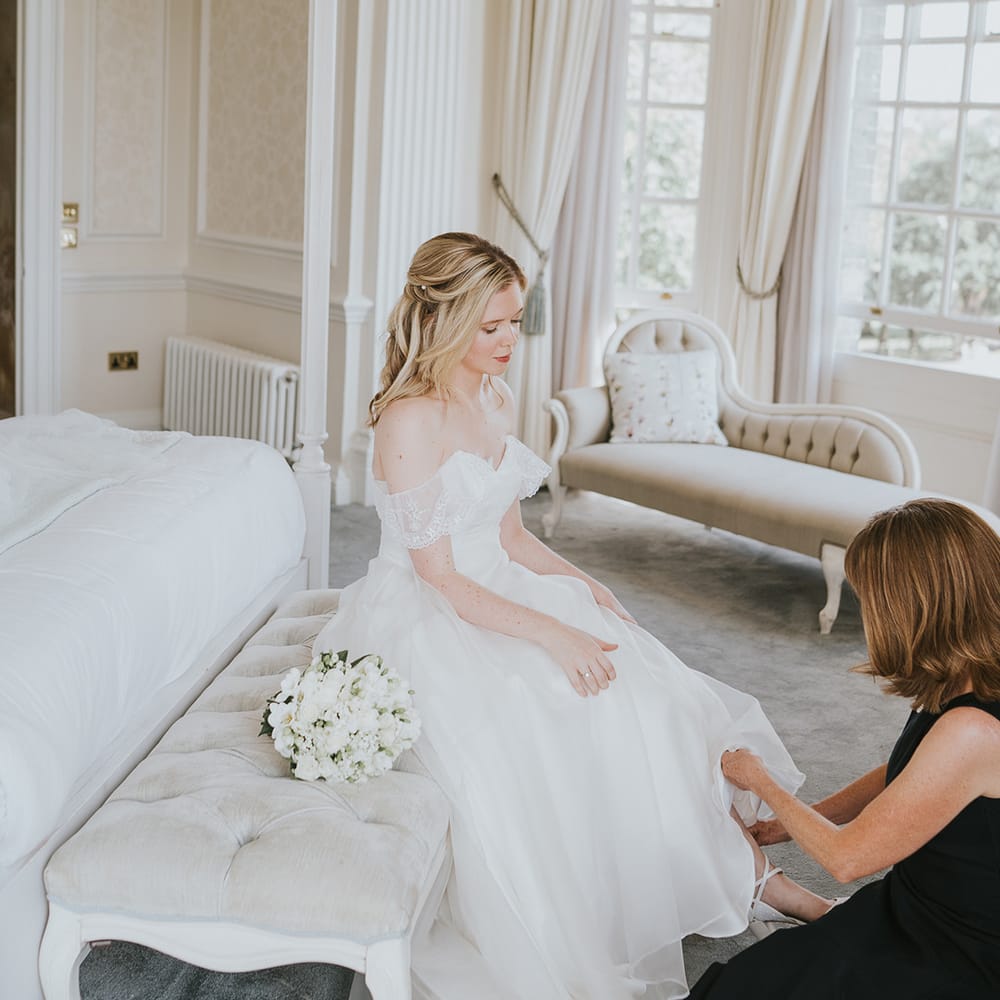 Bride in off the shoulder dress has white flower wedding bouquet gets help putting her shoes on for her wedding day 