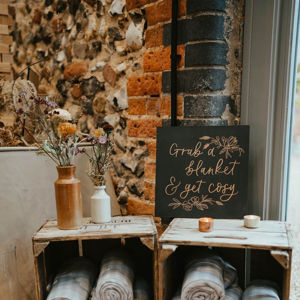 Wooden crates filled with blankets for wedding guests with tea lights, a black sign and autumnal flowers 