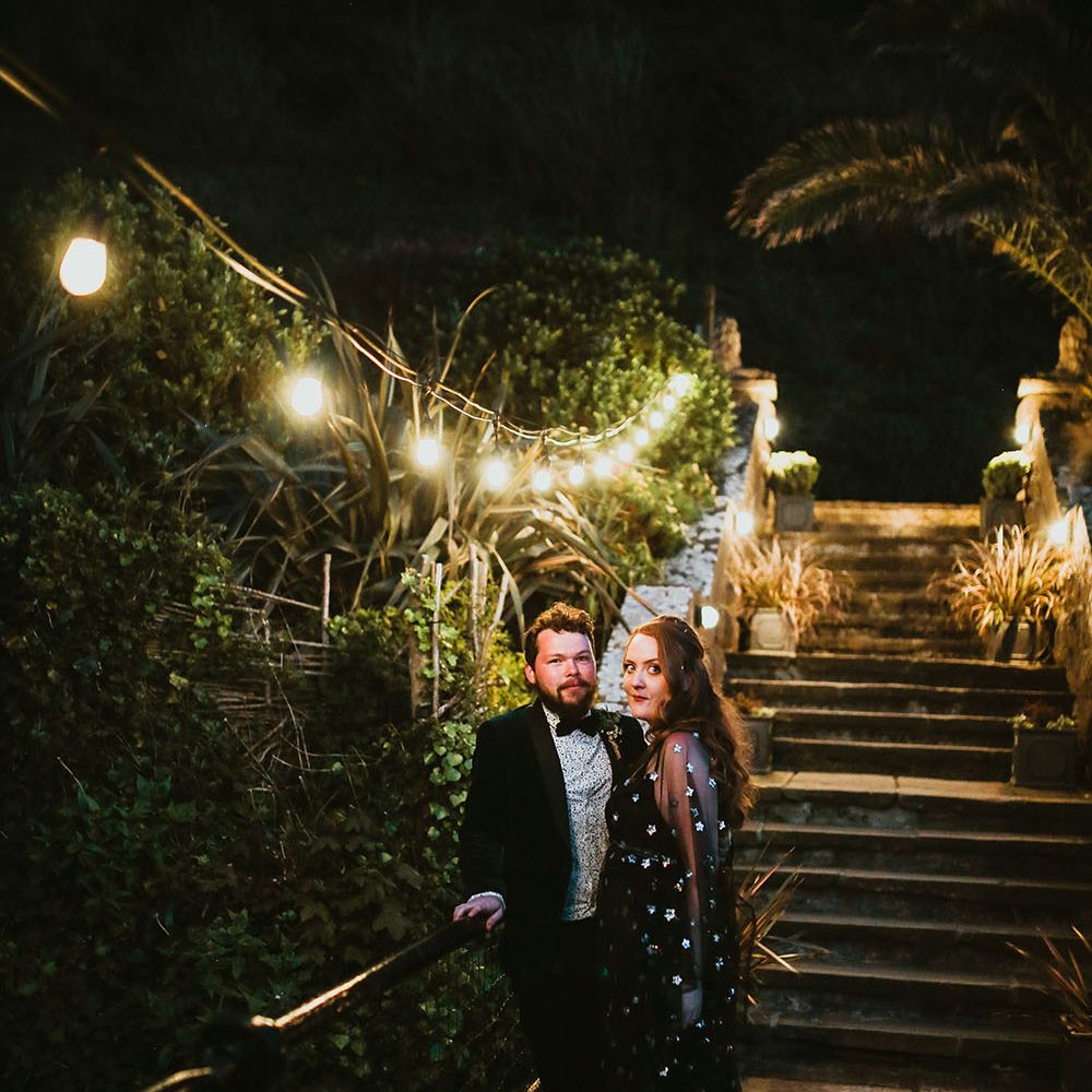 Bride wearing black celestial reception dress and groom in velvet dinner jacket stand beneath festoon lighting outdoors 