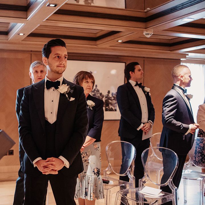 Groom stands in black tie for his wedding with a white flower buttonhole 