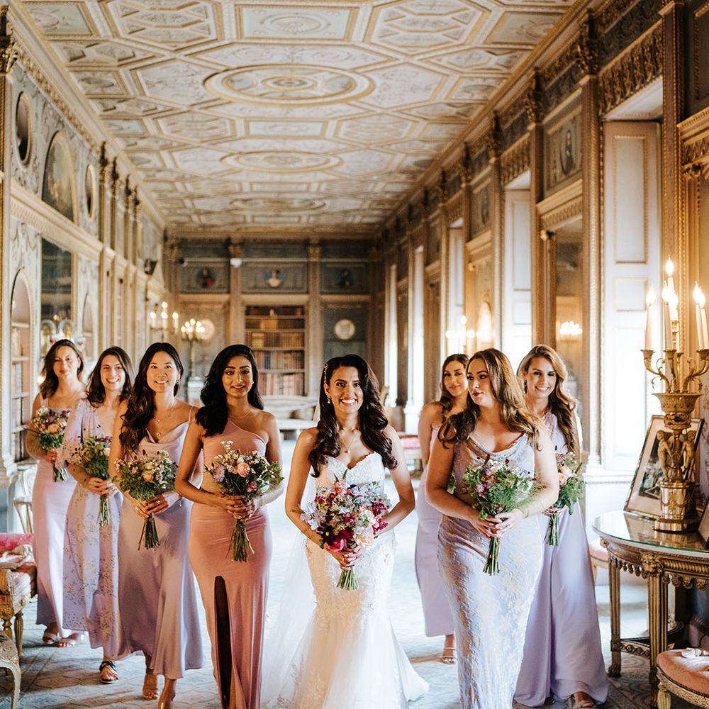 Bride walks through grand groom with her bridesmaids wearing lavender bridesmaid dresses in different shades and styles whilst holding brightly coloured floral bouquets 